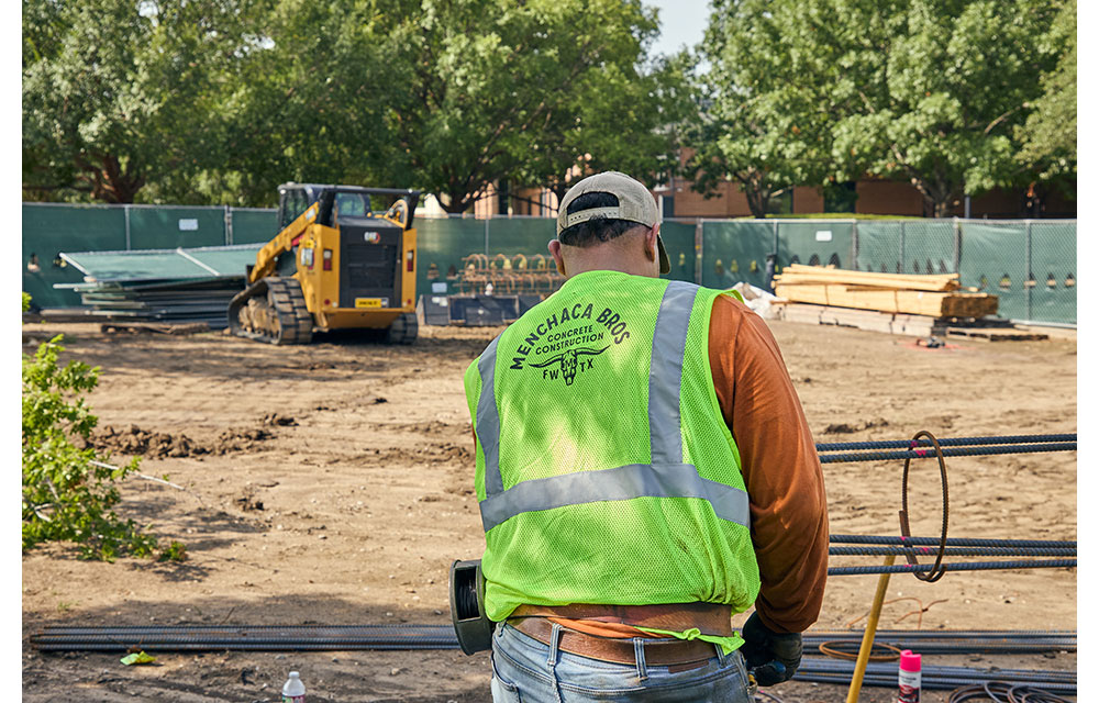 A construction worker wearing a neon safety vest labeled “Menchaca Bros Concrete Construction FWTX” stands at a job site with rebar in front of him and a yellow CAT skid steer in the background.